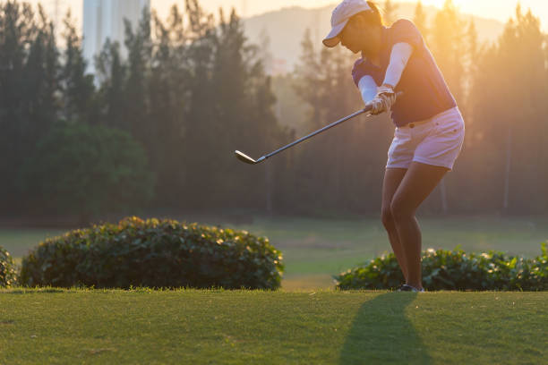 woman golf player in action of end downswing of wood driver, after hit the golf ball away from tee off to the fairway ahead, sunset scenery in background